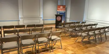 Empty conference room with rows of brown folding chairs facing a podium and banner at the front of the room.