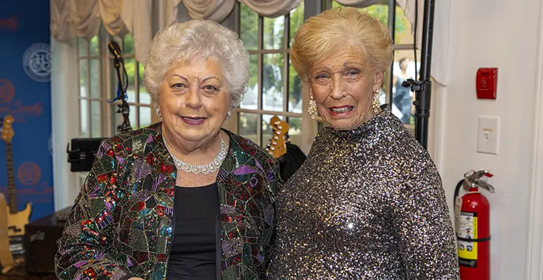 Two cheerful elderly women pose together backstage at a performance venue; one wears a colorful sequined jacket and the other a silver glitter top.