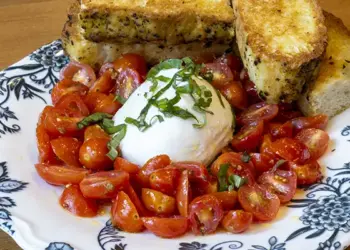 Plate of chopped cherry tomatoes around a burrata ball, topped with basil, with toasted bread slices on a blue-and-white patterned plate.