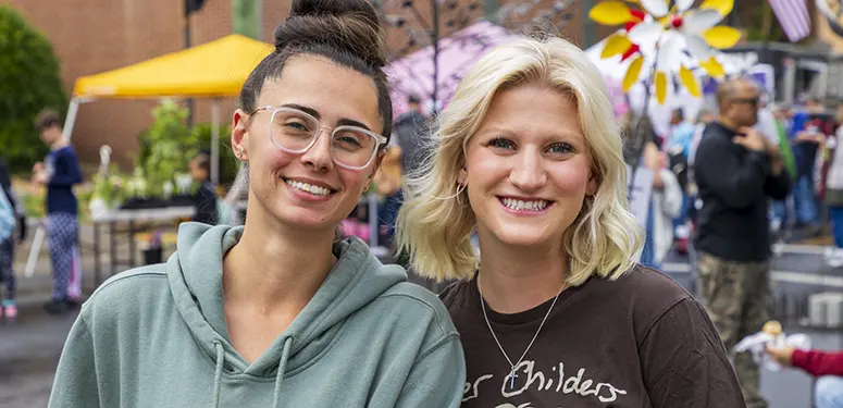 Two smiling women posing at an outdoor festival or market, with colorful tents and people in the background.