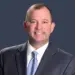 Smiling man in a dark suit and light blue tie against a dark blue background.