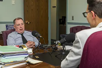 Chris Swam - Carroll News | Carroll Publications | Carroll County, MD Man in a gray sweater sits at a desk with a stack of papers, speaking into multiple microphones during an interview in a small studio.