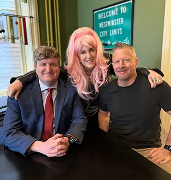 Dennis Sherri Randy - Carroll News | Carroll Publications | Carroll County, MD Three people sit at a table smiling; the middle person wears a pink wig and flanks two men in casual/business attire.