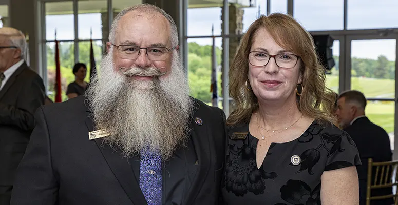Man with long white beard and glasses in a dark suit posing with a woman wearing glasses at a formal event or reception.