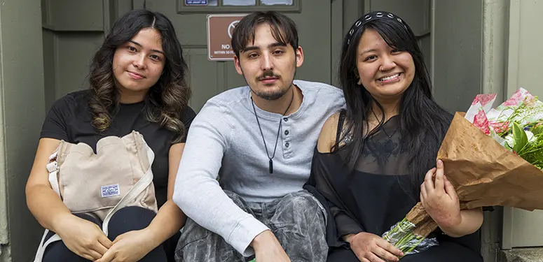 Three friends sit on outdoor steps posing for a photo; left woman holds a beige backpack, middle man wears a pendant, right woman smiles with a bouquet of flowers.