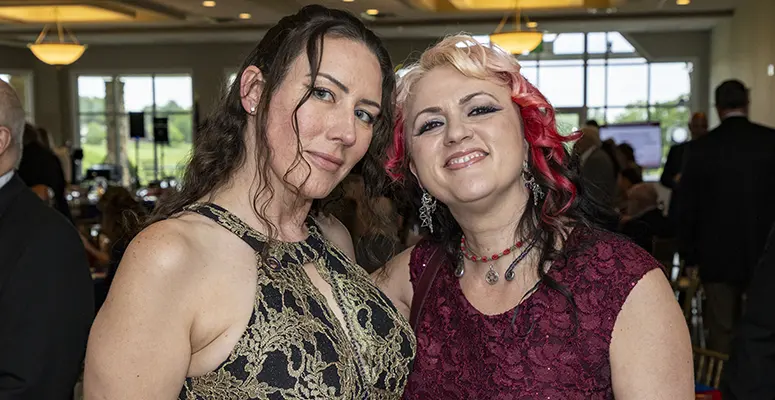 Two women in elegant dresses smiling and posing together at a formal event, indoors with guests in the background, warm lighting.