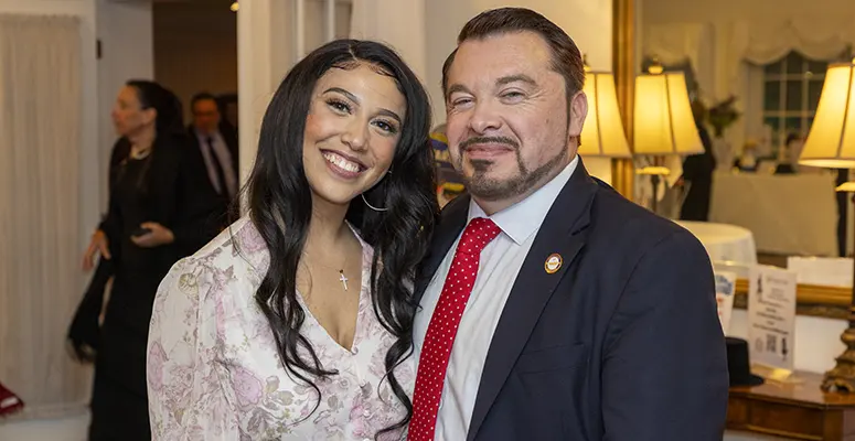 Smiling woman in a floral blouse and a man in a dark suit pose together at an indoor formal event with warm lighting in the background.