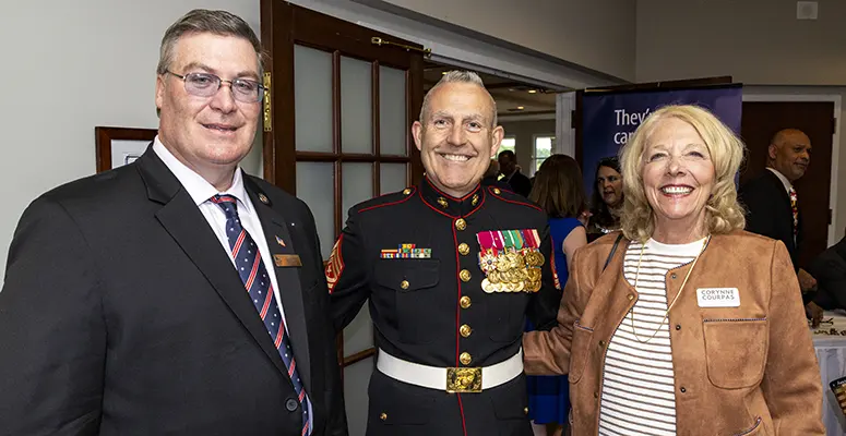 Three smiling adults at a formal event: a Marine in dress uniform with medals flanked by a man in a suit and a woman in a tan jacket nearby.