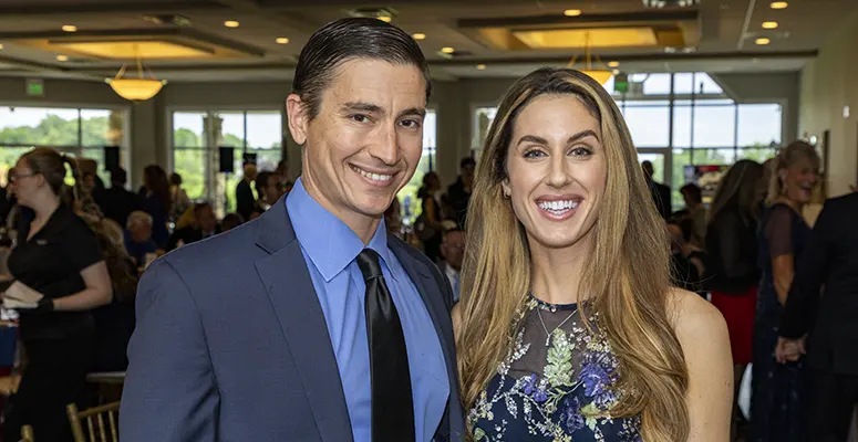 Smiling man in a blue suit and tie stands next to a woman in a floral dress at a social event indoors.