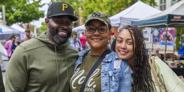 Three smiling friends posing at an outdoor market with tents in the background, wearing casual outfits and caps.