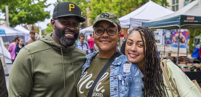Three smiling friends posing at an outdoor market with tents in the background, wearing casual outfits and caps.