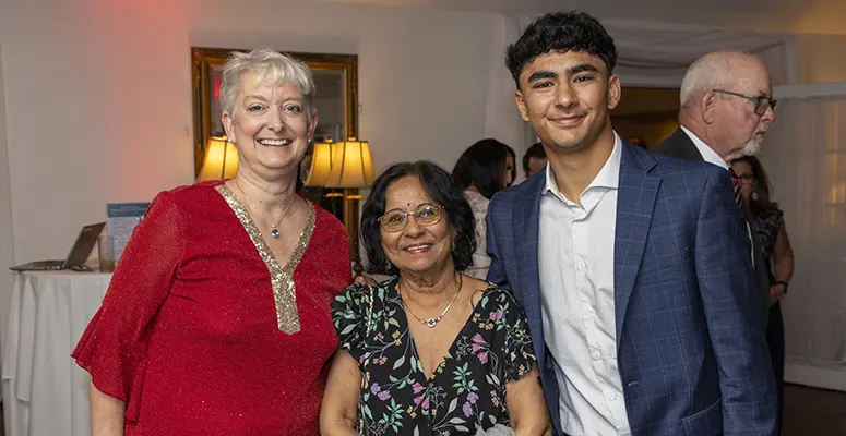 Three smiling people posing for a photo at a social event: an older woman in a red sequined top, a middle-aged woman in a floral dress, and a young man in a blue suit.