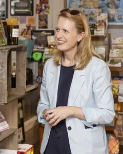 Woman in a light blue blazer smiles while standing in a colorful shop aisle filled with merchandise.