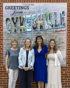 Four women pose in front of a brick wall mural that reads'Greetings from Sykesville, Maryland' with colorful letters.