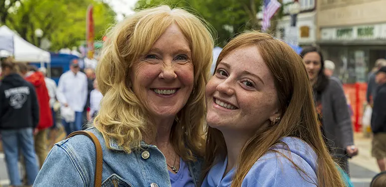 Two smiling women posing for a photo at an outdoor street festival with tents and shoppers in the background, hugging together.