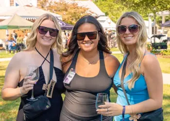 Three women wearing sunglasses pose together at an outdoor festival, each holding a wine glass.