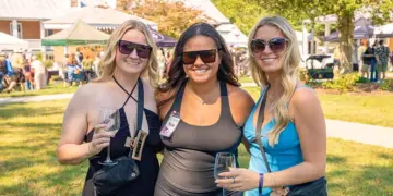 Three women wearing sunglasses pose together at an outdoor festival, each holding a wine glass.