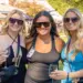 Three women wearing sunglasses pose together at an outdoor festival, each holding a wine glass.