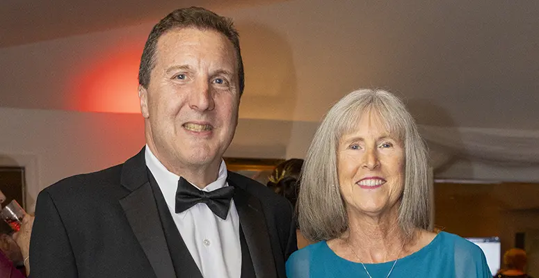 Smiling man in a black tuxedo with bow tie standing next to a woman in a teal dress at a formal event.