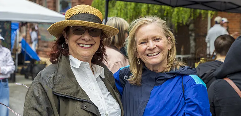 Two smiling women pose together at an outdoor event, one wearing a straw hat and glasses, the other in a blue jacket.