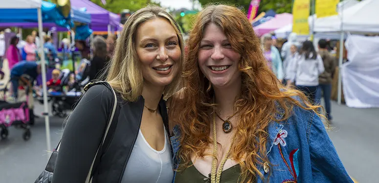 Two women smile at the camera while posing together at an outdoor street market with colorful tents in the background.