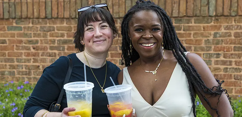 Two smiling women stand together outdoors, each holding a clear cup with fruit slices and straws in front of a brick wall.