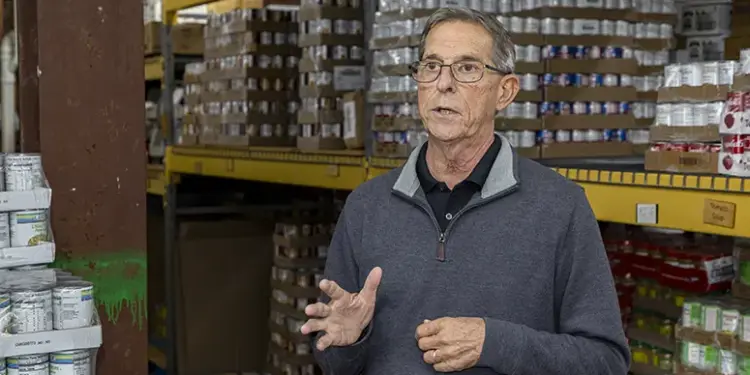 Older man wearing glasses speaks and gesticulates in a warehouse aisle with shelves of boxed goods behind him, suggesting a briefing or tour.
