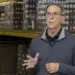 Older man wearing glasses speaks and gesticulates in a warehouse aisle with shelves of boxed goods behind him, suggesting a briefing or tour.