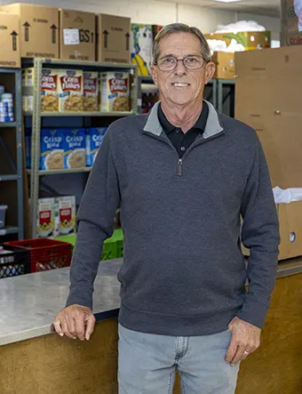 Smiling older man with glasses behind a counter in a food pantry; shelves of cereal and cardboard boxes in the background.