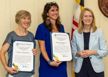 Three women stand indoors, two in blue-toned dresses holding framed certificates and smiling as the third woman in a light blue blazer stands beside them.