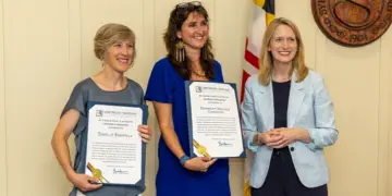 Three women stand indoors, two in blue-toned dresses holding framed certificates and smiling as the third woman in a light blue blazer stands beside them.