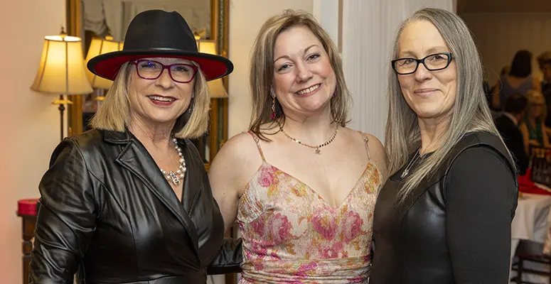 Three smiling women posing together indoors at a social event; one in a black leather jacket and red-trim hat, another in a pink floral dress, and the third in a black top with glasses.