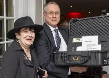 Two smiling adults pose at an event, with a man holding an open hard-shell equipment case containing papers and items for auction or raffle in a banquet setting.