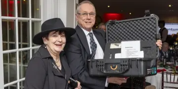 Two smiling adults pose at an event, with a man holding an open hard-shell equipment case containing papers and items for auction or raffle in a banquet setting.