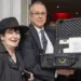 Two smiling adults pose at an event, with a man holding an open hard-shell equipment case containing papers and items for auction or raffle in a banquet setting.
