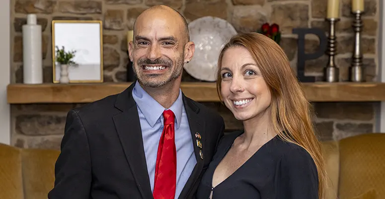 Smiling man in a black suit and red tie stands beside a woman with long red hair in a cozy living room with a stone fireplace behind them.