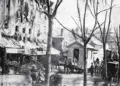 Historic city street with horse-drawn carriages, vendors, and pedestrians along a row of shops on a tree-lined avenue.