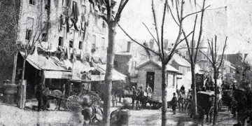 Historic city street with horse-drawn carriages, vendors, and pedestrians along a row of shops on a tree-lined avenue.