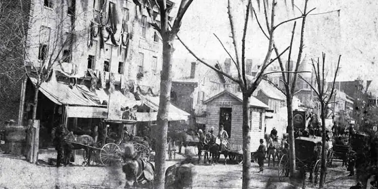 Historic city street with horse-drawn carriages, vendors, and pedestrians along a row of shops on a tree-lined avenue.