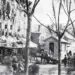 Historic city street with horse-drawn carriages, vendors, and pedestrians along a row of shops on a tree-lined avenue.