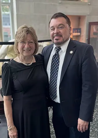 Two smiling adults pose together indoors, the woman in a black dress and the man in a dark suit and patterned tie.