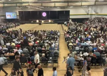 Large school gym packed with hundreds of attendees seated and facing a stage with screens on either side.