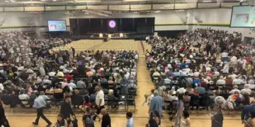 Large school gym packed with hundreds of attendees seated and facing a stage with screens on either side.