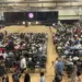 Large school gym packed with hundreds of attendees seated and facing a stage with screens on either side.