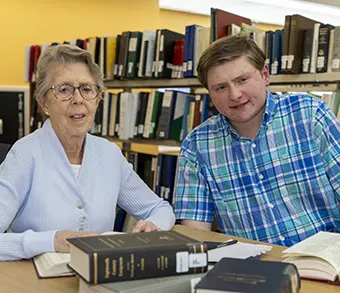 An elderly woman wearing a light cardigan and glasses sits at a library table with a younger man; they study surrounded by books.