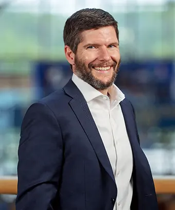 Professional headshot of a smiling man with dark hair and beard, wearing a navy suit and white shirt, in an office setting.