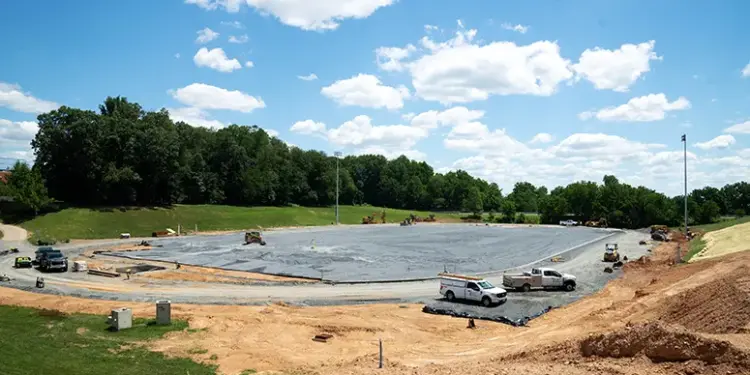 Construction site for a sports field in progress: a large excavated area lined with black tarp, with several trucks and equipment and dirt mounds around it, trees in the background.