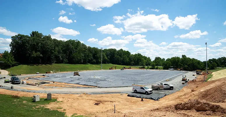 Construction site for a sports field in progress: a large excavated area lined with black tarp, with several trucks and equipment and dirt mounds around it, trees in the background.