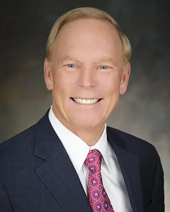 Head-and-shoulders portrait of a smiling man in a navy suit and pink patterned tie, against a blurred dark background.