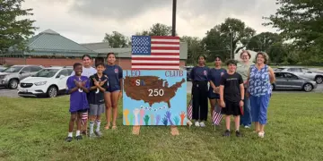Group of children and adults posing beside a handmade sign reading LEO Club USA 250, with an American flag overhead.